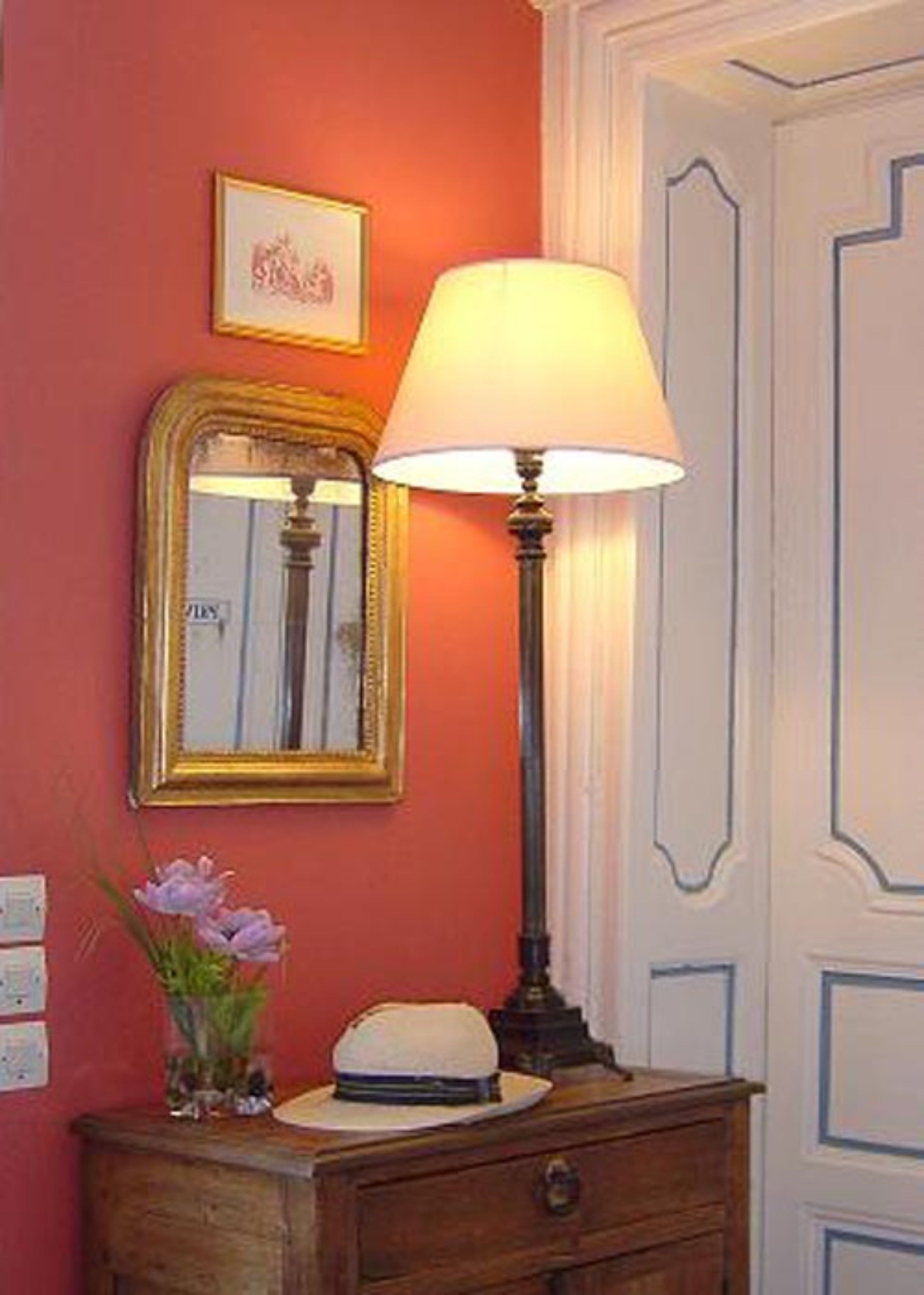 Une pièce avec un miroir, une lampe, une commode et un vase rempli de fleurs.