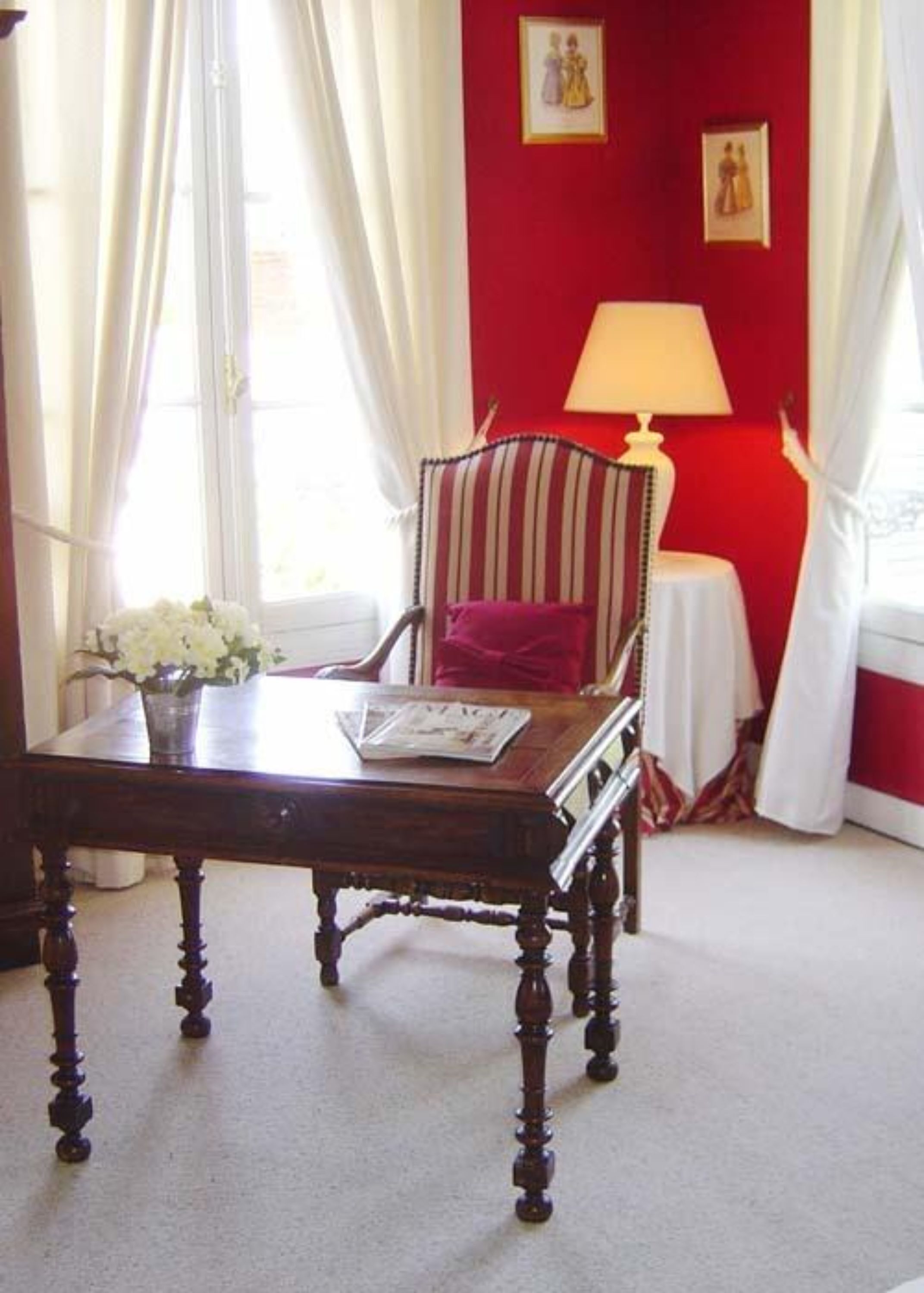Una habitación con una mesa de madera, un sillón a rayas, una mesita con una lámpara y un ventanal con cortinas.