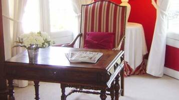 Una habitación con una mesa de madera, un sillón a rayas, una mesita con una lámpara y un ventanal con cortinas.