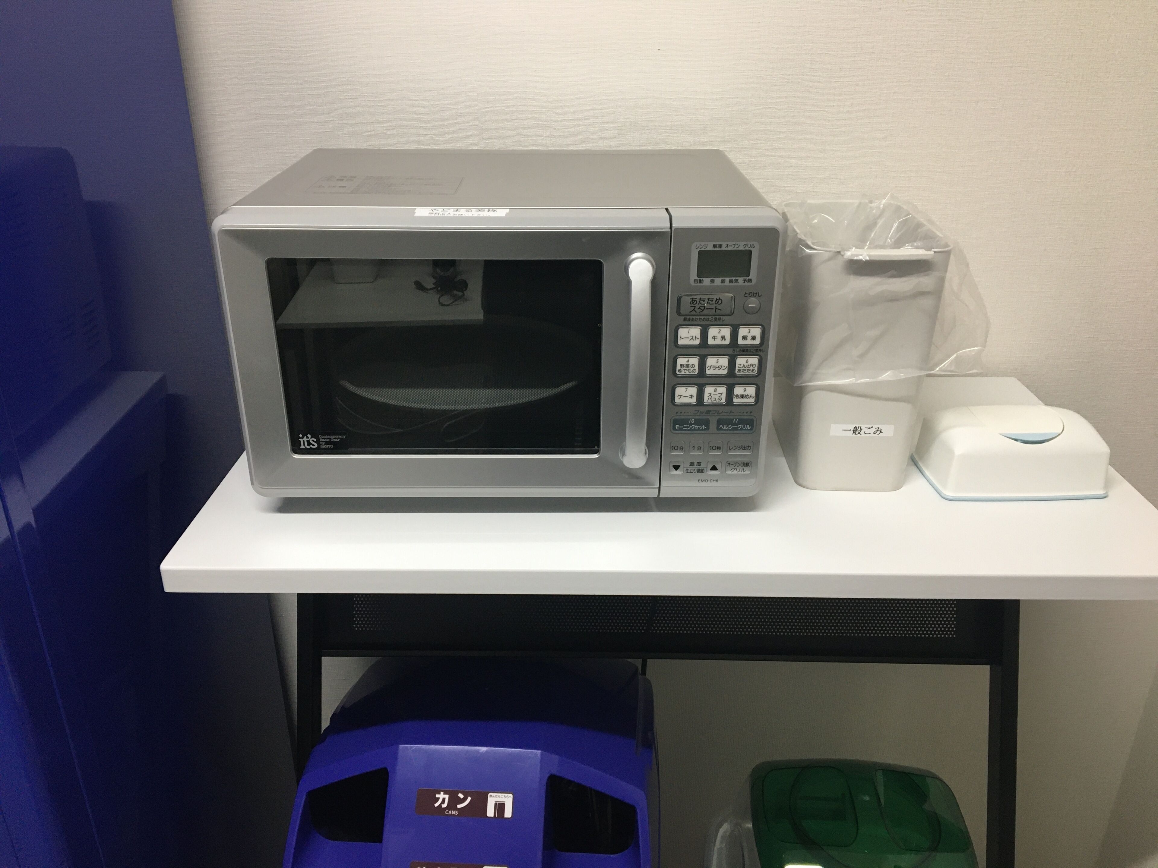 Un microondas sobre una mesa blanca, con una bolsa de plástico y un electrodoméstico blanco a su lado.