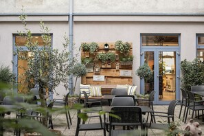An outdoor seating area with tables and chairs, a wooden wall display with plants and birdhouses, and a building entrance with a blue door. - myNext - Hotel Rudy (Vienna)