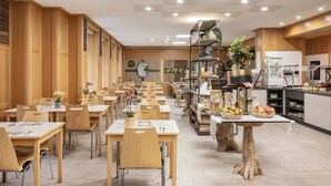 A spacious dining area with multiple tables and chairs, a central serving table with fruits, and a kitchen area in the background. - myNext - Hotel Rudy (Vienna)
