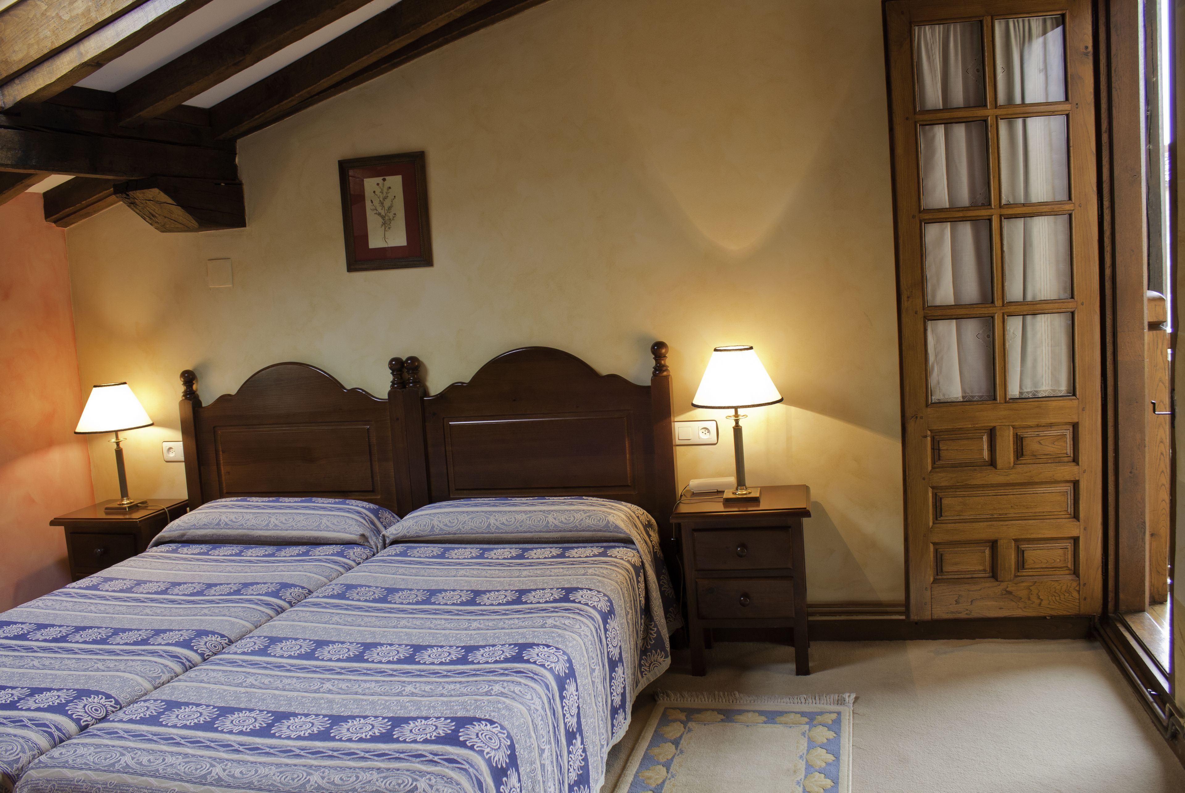 Un dormitorio con dos camas, cabeceros de madera, lámparas de noche, un cuadro enmarcado en la pared y una puerta de madera con paneles de vidrio.