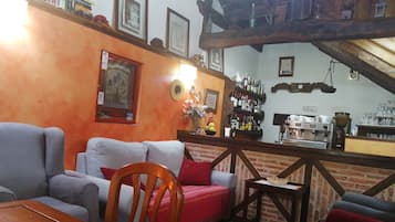 Un saló d'estar acollidor amb una taula baixa de fusta, un sofà i un butaca vermella. Una paret de maó amb una estanteria de llibres i quadres emmarcats. Una màquina de cafè i una taula petita amb un cistó es veuen al fons.