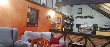 Un salón acogedor con una mesa de centro de madera, un sofá y un sillón rojo. Se observa una pared de ladrillo con una estantería llena de libros y cuadros enmarcados. En el fondo, una cafetera y una pequeña mesa con una cesta.