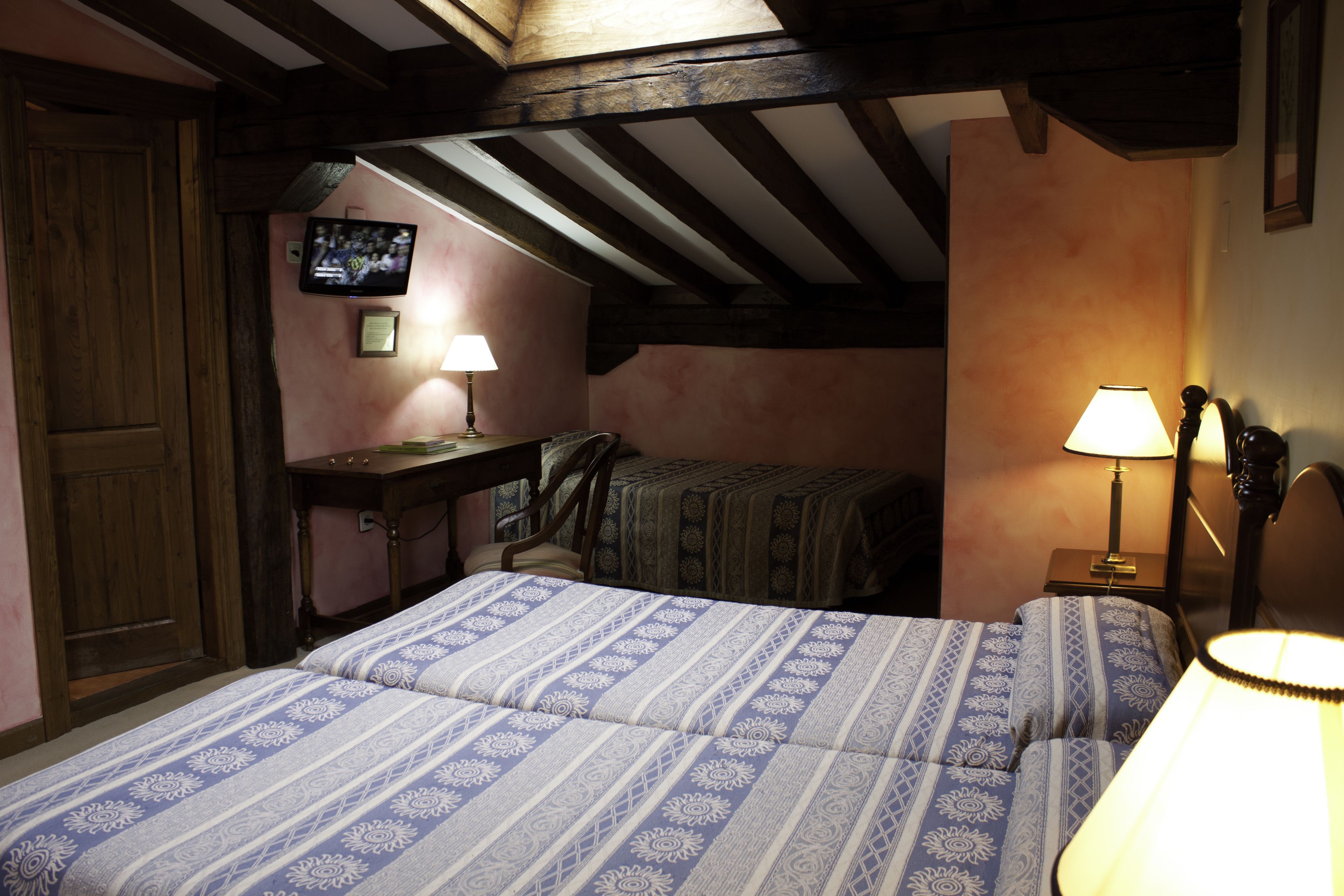 A bedroom with a wooden ceiling, a bed with patterned bedding, a desk with a lamp, and a television on the wall.