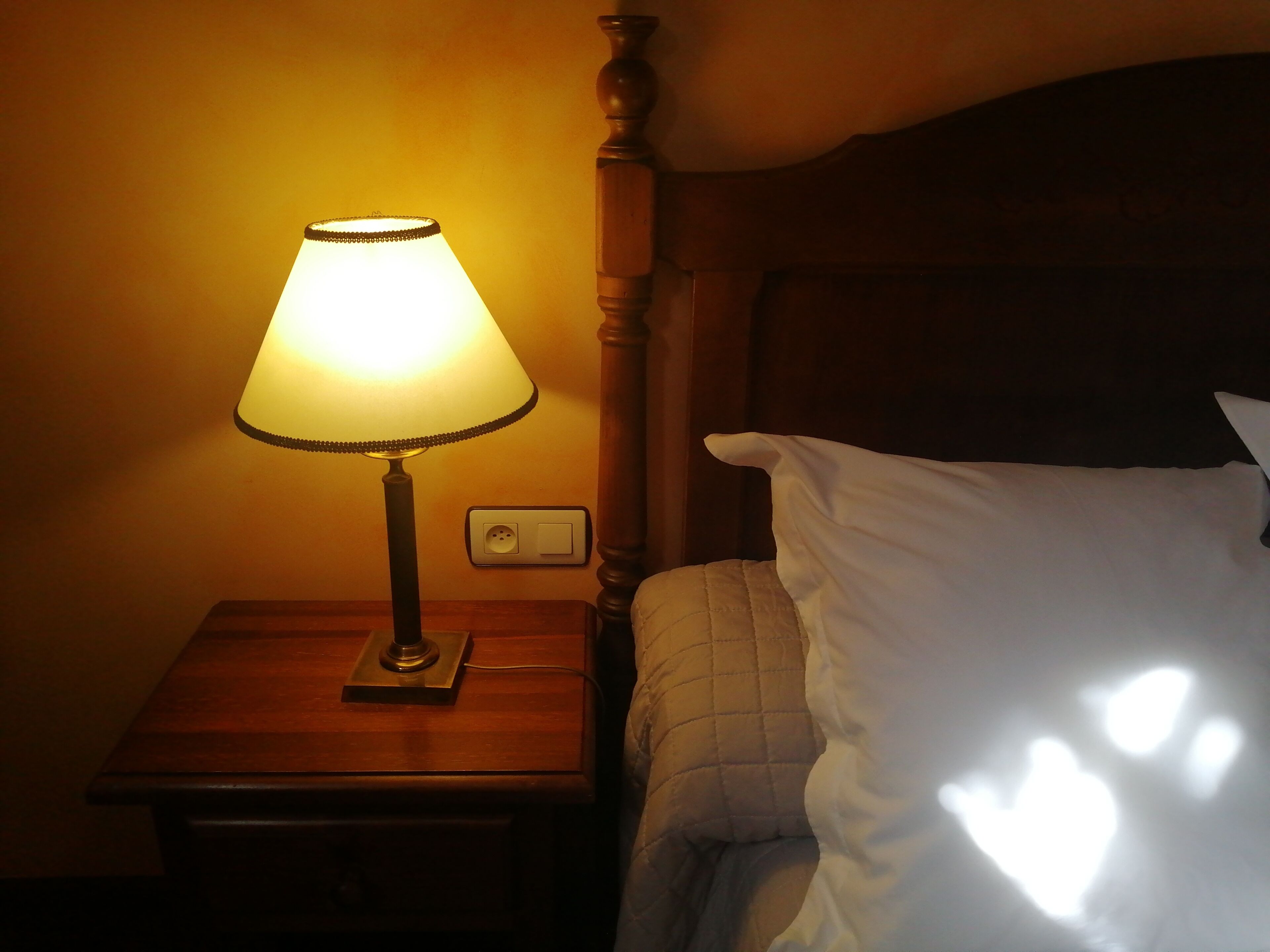 A bedside table with a lamp, a power outlet, and a bed with a white pillow.