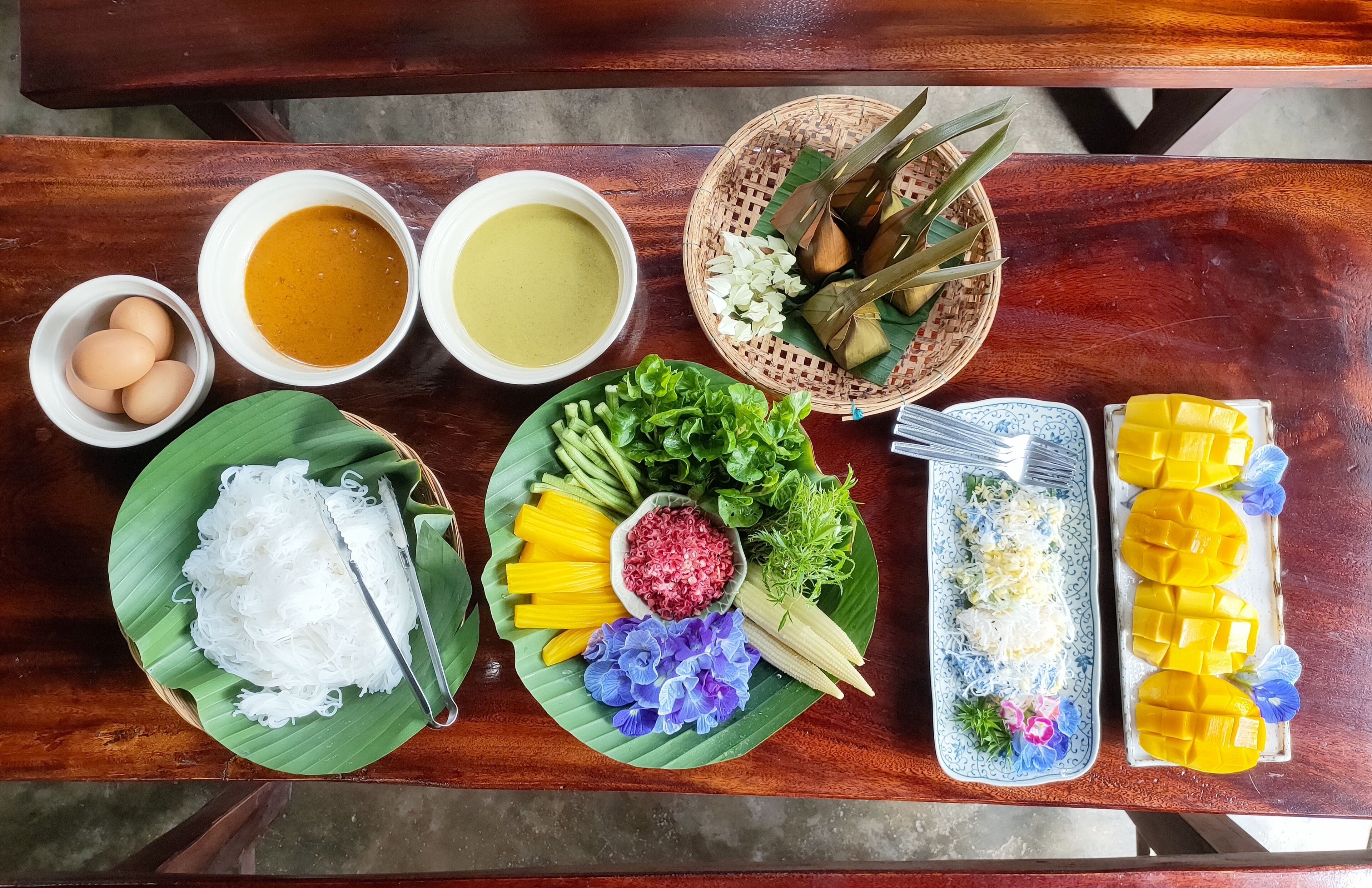 A wooden table with various dishes including noodles, eggs, and mango slices.