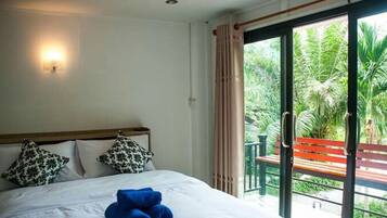 Een hotelkamer met een bed, twee bedkussens met dessin, een blauwe handdoek, een wandlamp en een schuifdeur naar een buitenruimte met een bankje en beplanting.