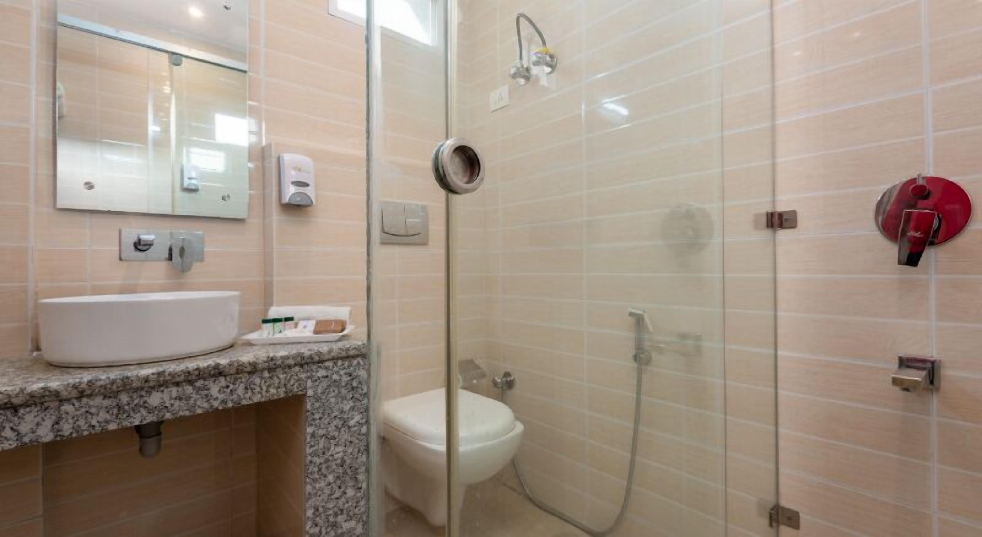 Un baño con encimera de granito, un lavamanos blanco, un espejo, un inodoro y una zona de ducha con mampara de vidrio y cabezal de ducha manual.