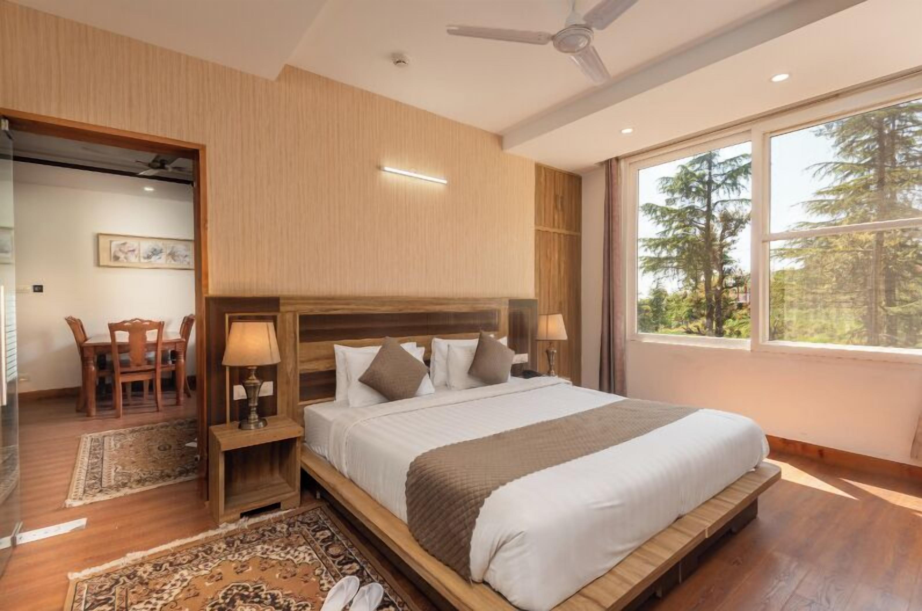 Ein Schlafzimmer mit einem großen Bett, einem Holz-Kopfteil, Nachttischlampen, einem Nachttisch, einem Schreibtisch mit Stuhl und einem großen Fenster mit Blick auf Bäume.