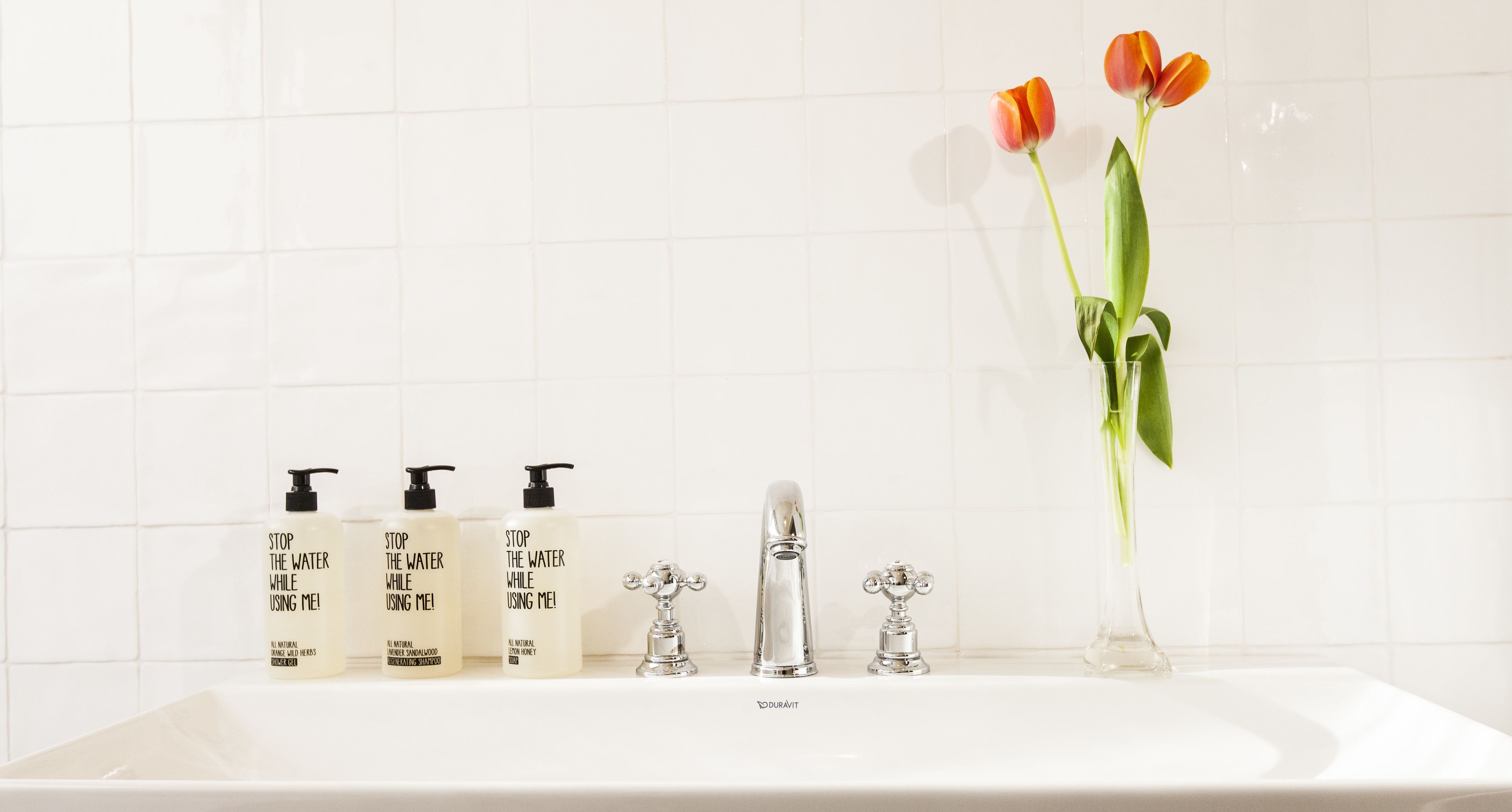 A bathroom sink with three bottles of 'Stop the Water While Using Me!' shampoo, a silver faucet, and a vase with two tulips.