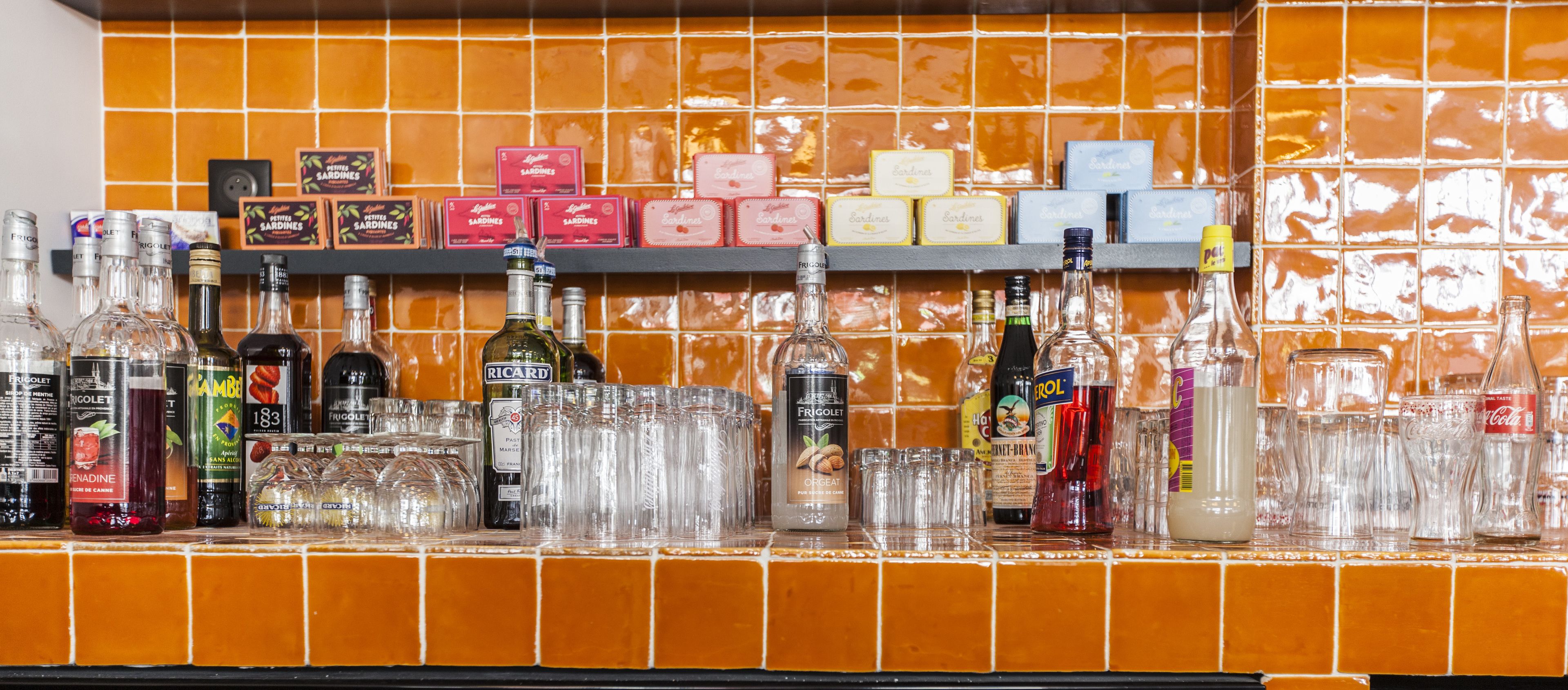A bar area with various bottles and glasses, shelves with boxes of snacks, and a tiled backsplash.