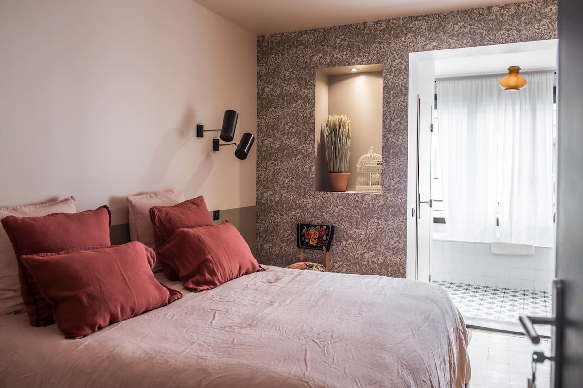 Un dormitorio con cama, almohadas, una lámpara de pared, una planta en maceta y una puerta con cortinas.