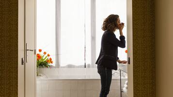 A woman standing in a bathroom with a shower curtain, a sink, and a window.