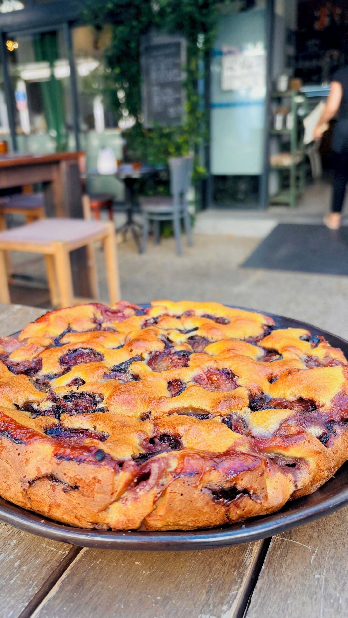 A round fruit tart on a metal plate, with a blurred outdoor seating area in the background.