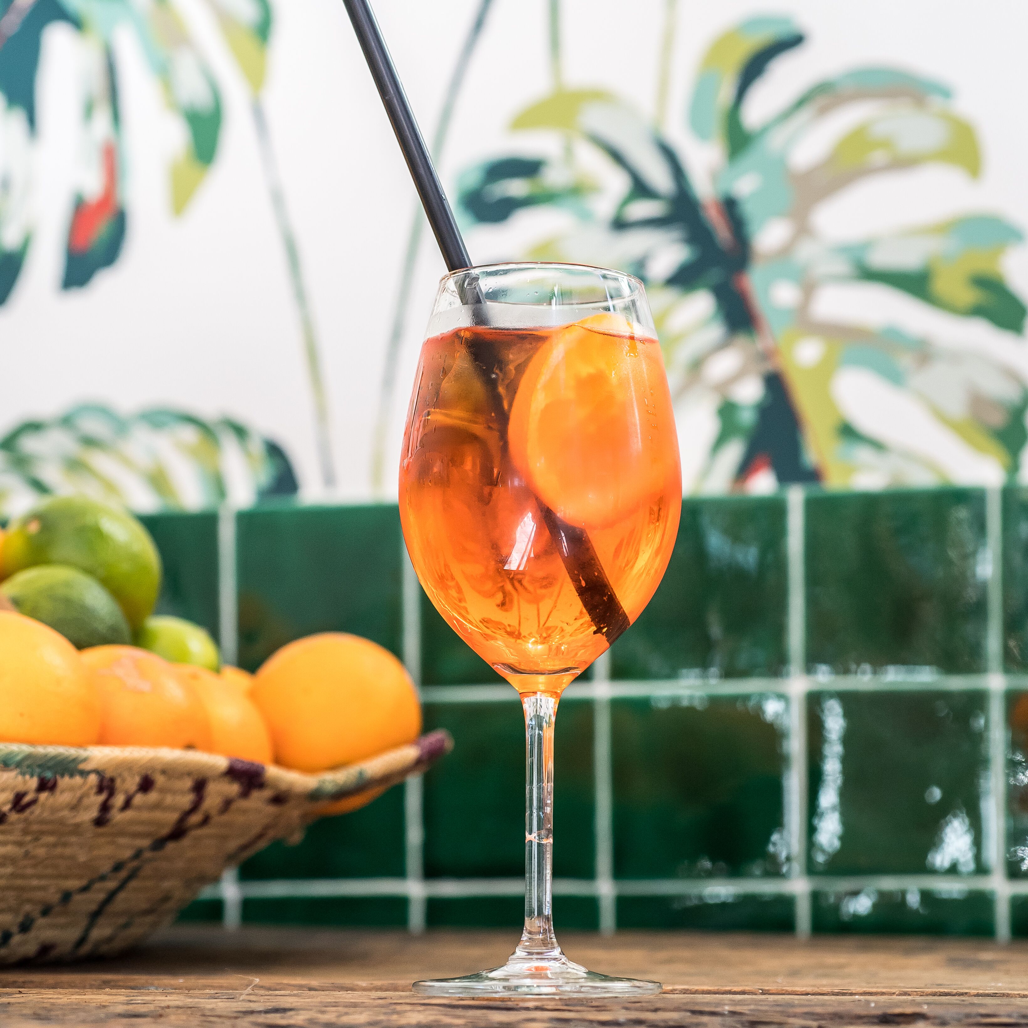 A glass of orange-colored drink with a slice of orange and a straw, placed on a wooden surface with a green tiled wall and a bowl of oranges in the background.