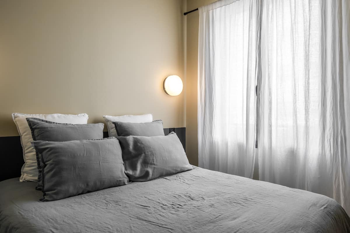 Una cama bien hecha con ropa de cama gris, una lámpara de pared redonda y cortinas translúcidas.