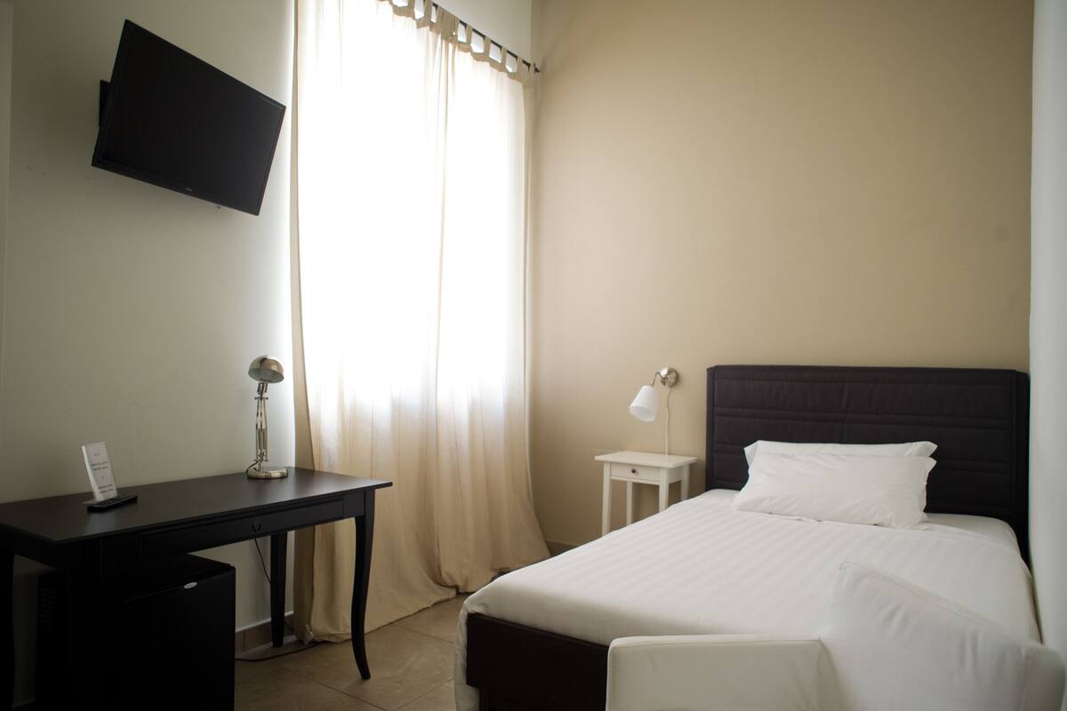 A hotel room with a bed, a desk, a chair, a TV mounted on the wall, and a window with curtains.