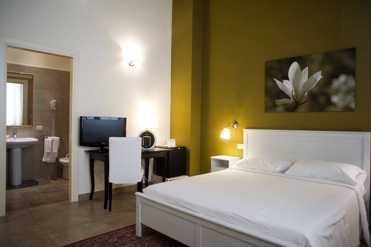 A hotel room with a bed, a desk with a computer, a TV, a bathroom with a sink and toilet, and a painting on the wall.