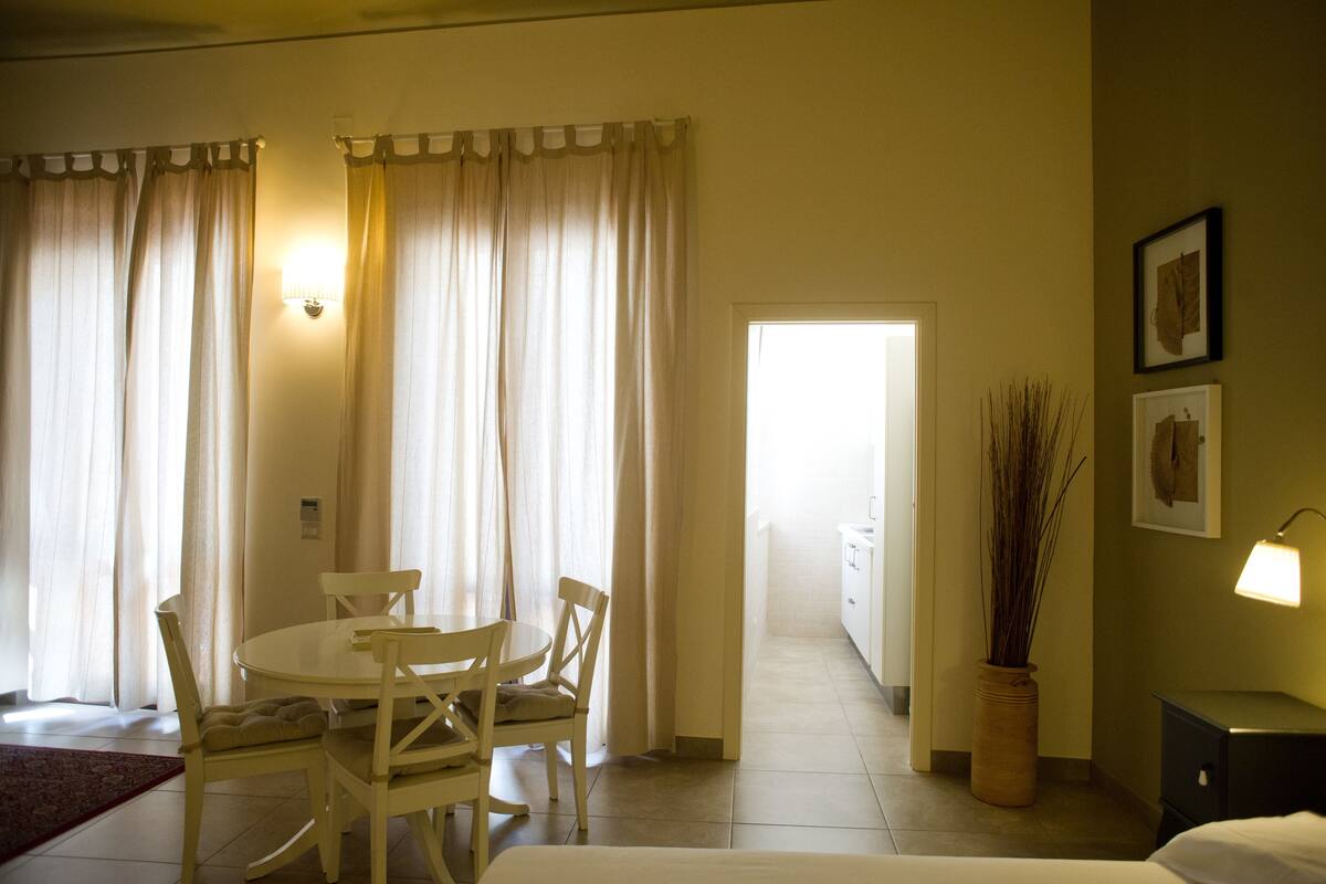 A hotel room with a dining table and chairs, a bed, a nightstand, a lamp, and a potted plant.