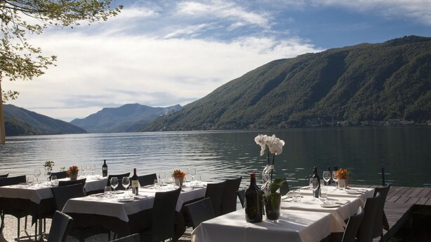 A lakeside restaurant with tables set for dining, overlooking a lake and mountains.