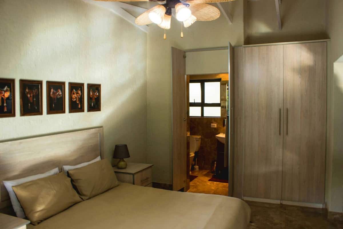 A bedroom with a bed, a nightstand, a lamp, a wardrobe, and a bathroom visible through an open door.
