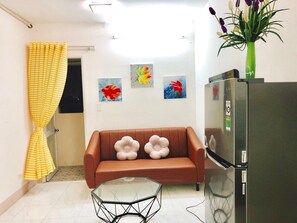 Una sala de estar con un sofá marrón, dos cojines con forma de flor, una mesa de centro de vidrio, un refrigerador y un jarrón con flores encima.