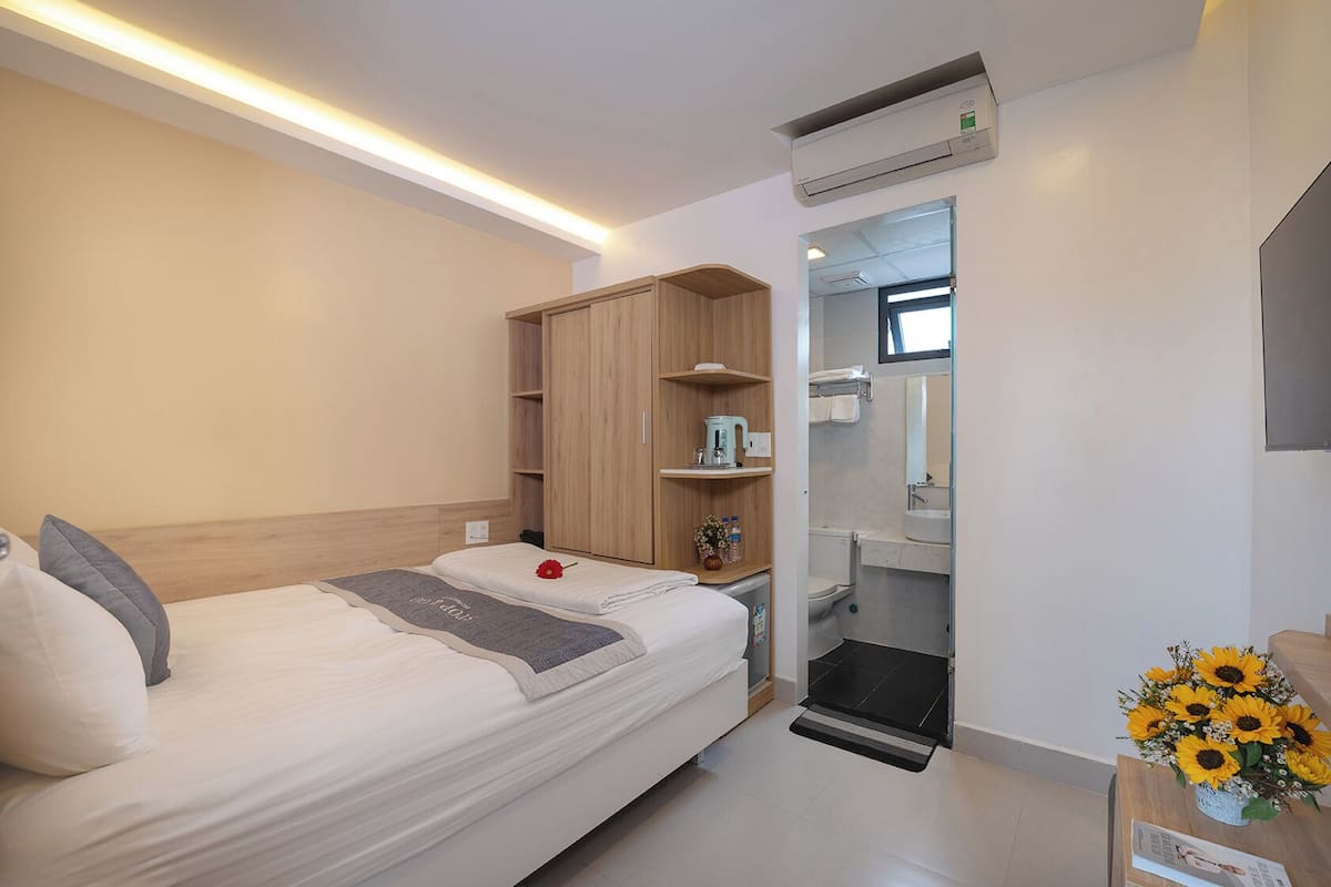 A modern hotel room with a neatly made bed, a wooden wardrobe, a small kitchenette, and a bathroom visible through an open door.