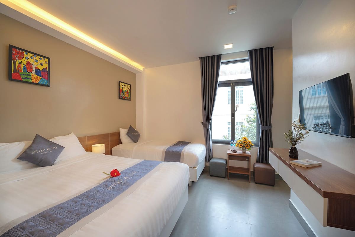 A hotel room with two beds, a desk with a vase of flowers, a TV on the wall, and a window with curtains.