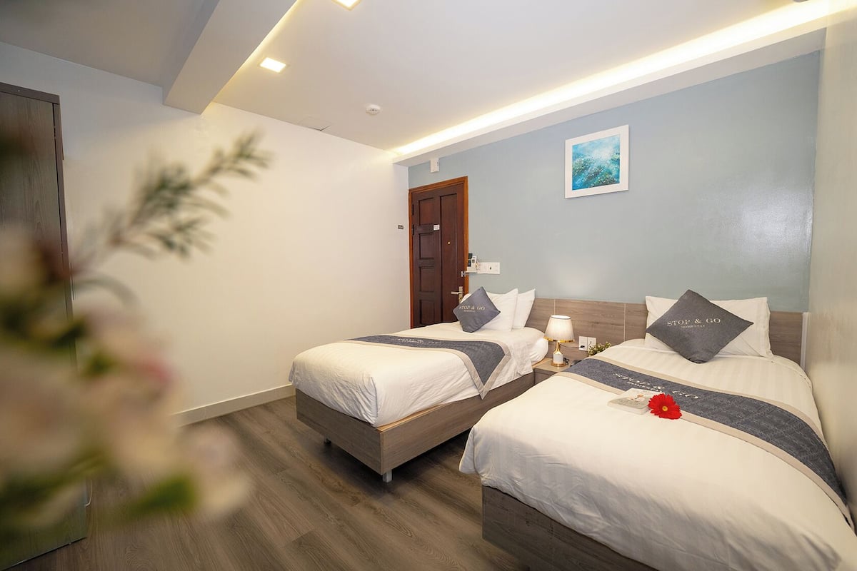 A hotel room with two beds, a wooden floor, a door, a wall-mounted light, and a small painting.