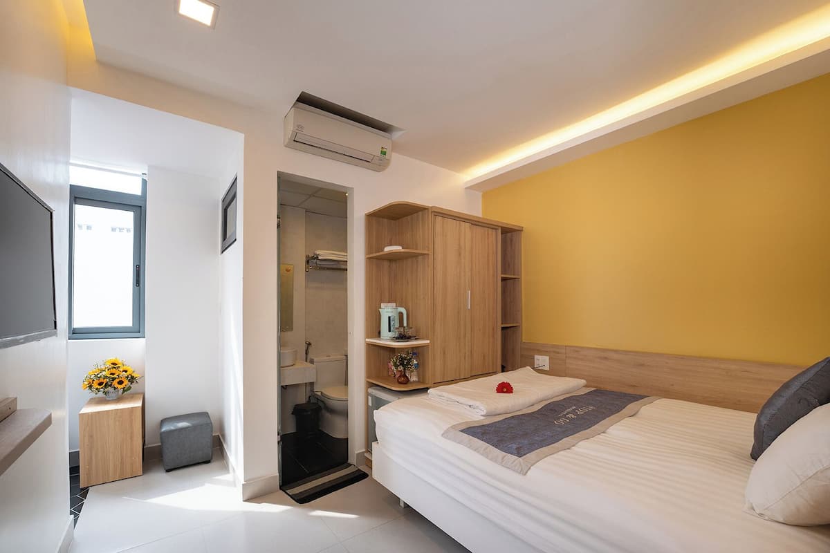 A bedroom with a bed, a wardrobe, a small table with a vase of flowers, a window, and an air conditioning unit.