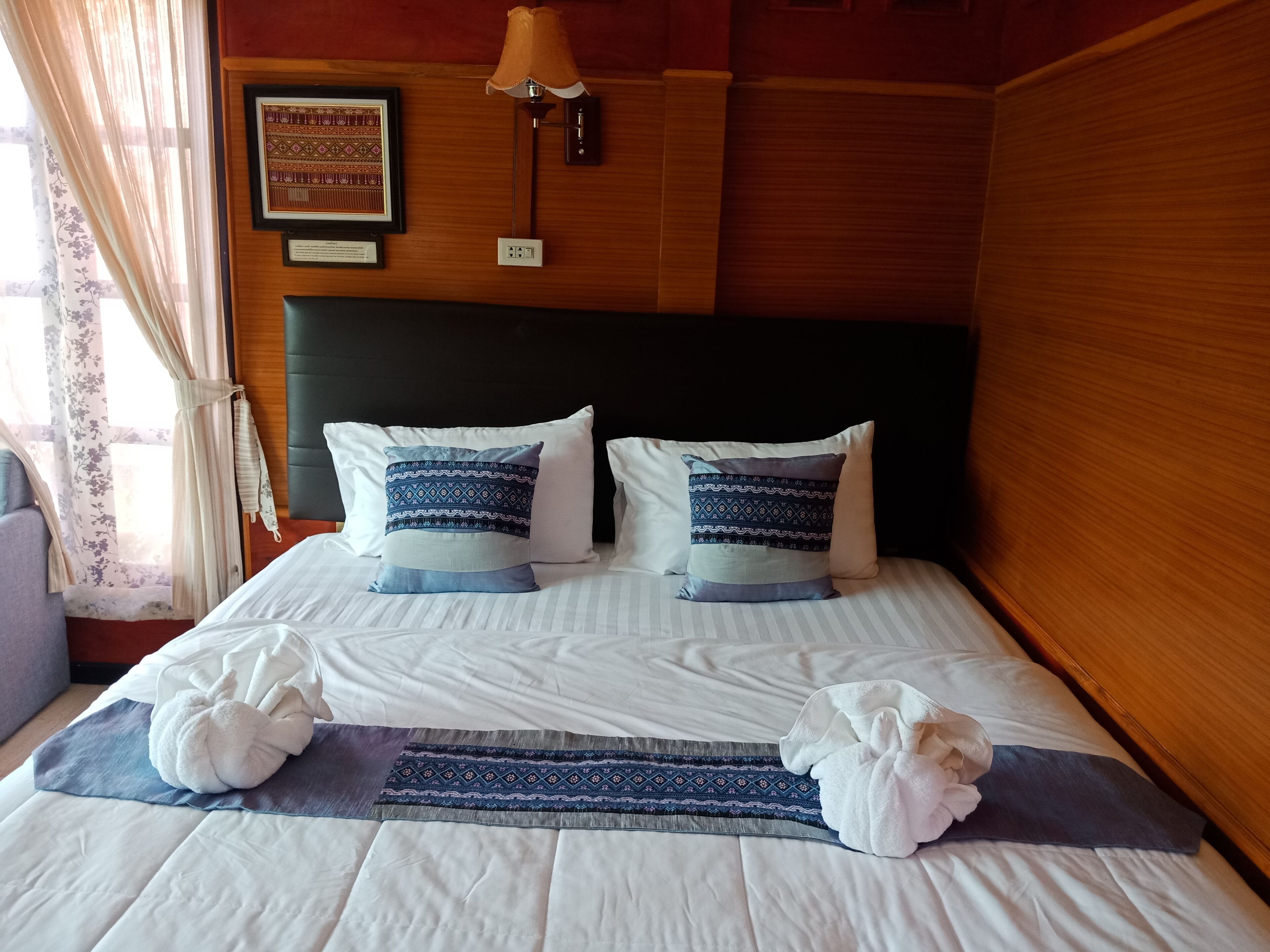 A neatly made bed with white linens, two decorative pillows with a blue pattern, and two rolled towels on a striped bedspread.