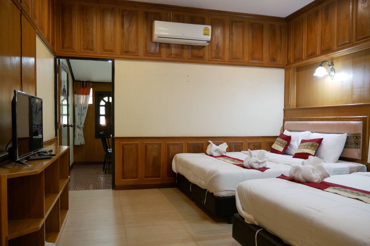 Una habitación de hotel con paneles de madera, dos camas con sábanas blancas y almohadas decorativas, un televisor de pantalla plana sobre un soporte de madera y un equipo de aire acondicionado montado en la pared.