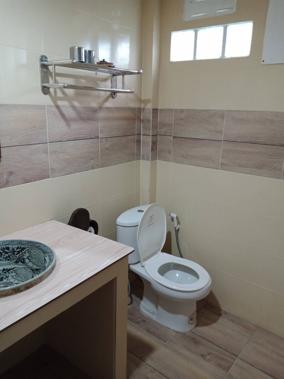 Casa de banho com sanita, lavatório com uma tigela decorativa, prateleira com artigos de higiene pessoal e janela que proporciona luz natural.