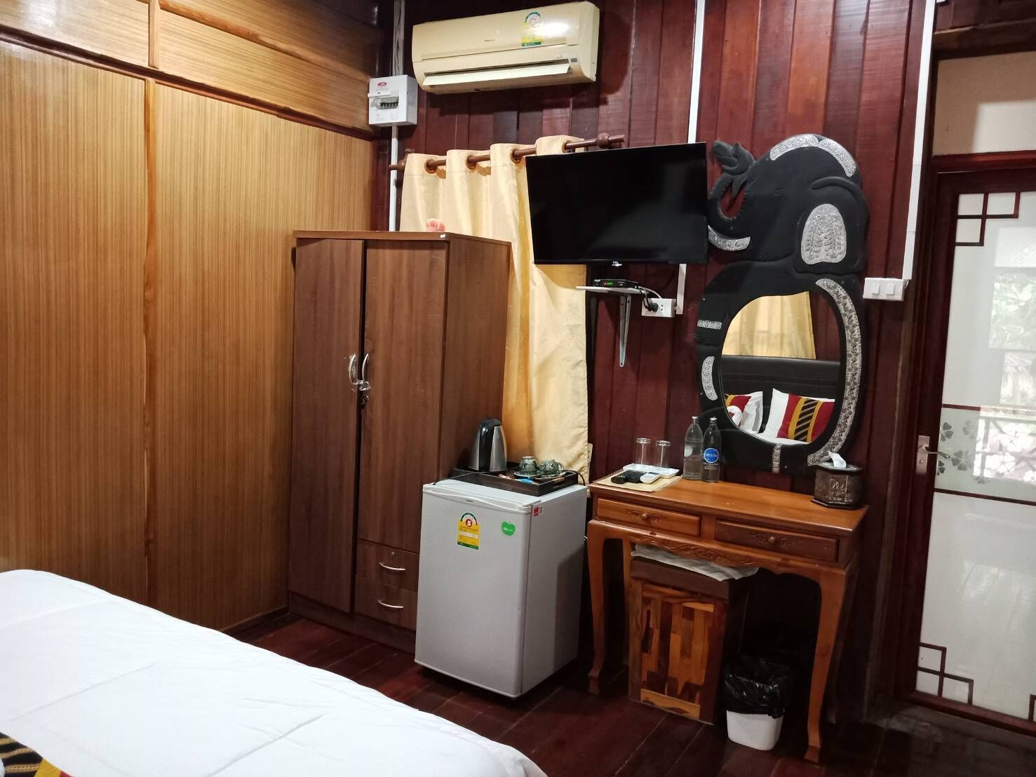 Ένα δωμάτιο ξενοδοχείου με ξύλινη επένδυση στους τοίχους, ένα κρεβάτι, ένα μικρό ψυγείο, ένα γραφείο με καθρέφτη και μια τηλεόραση τοποθετημένη στον τοίχο.