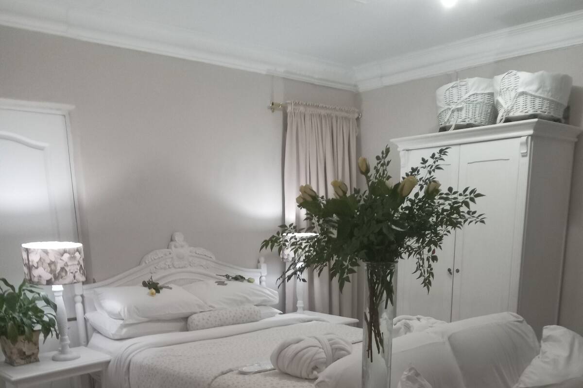 A white-themed bedroom with a bed, bedside tables, a nightstand, a wardrobe, and a vase with flowers.