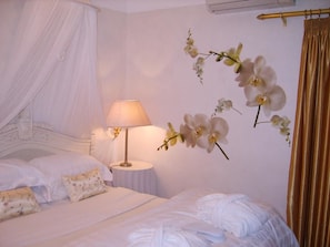Quarto com cama de dossel, luminária de cabeceira e arte de parede com orquídeas.