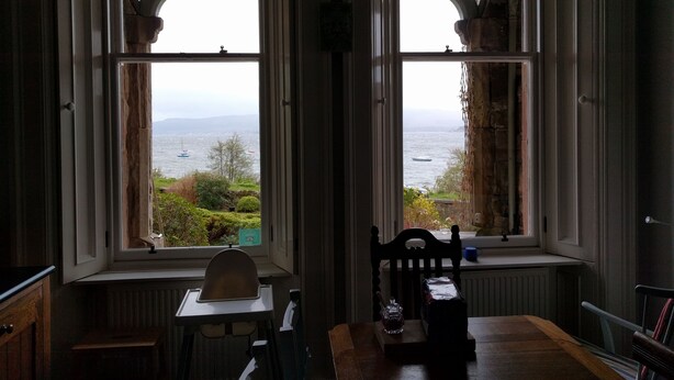 A room with two large windows overlooking a body of water, a wooden table, a chair, and a radiator. - Cragowlet House East (Helensburgh)