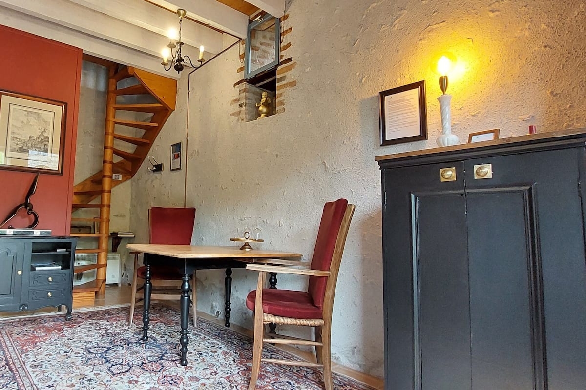 A room with a wooden staircase, a dining table with chairs, a cabinet, and a patterned rug.