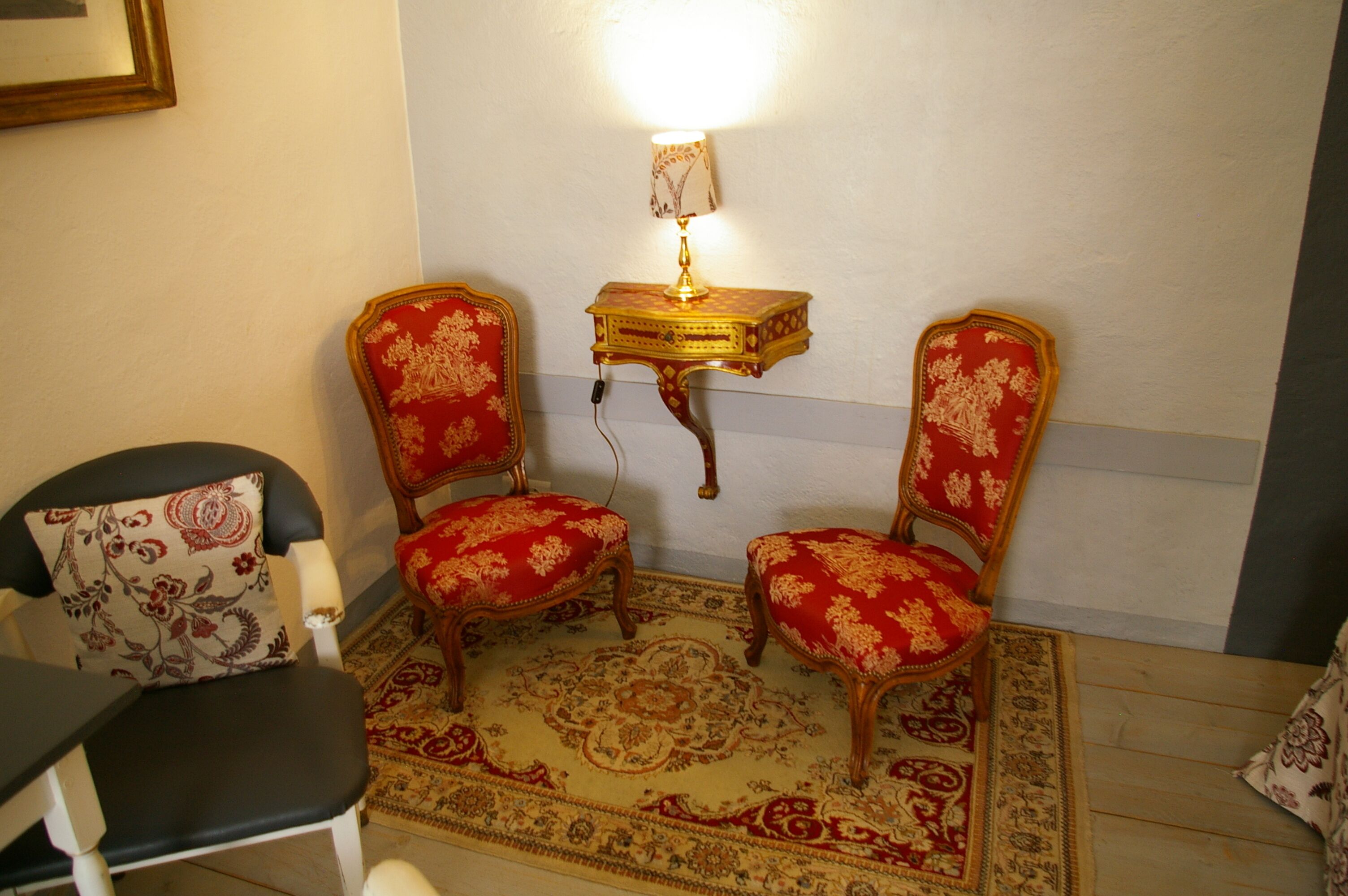 A room with two ornate red upholstered chairs, a small table with a lamp, and a patterned rug.