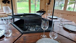 A wooden dining table with a built-in grill, surrounded by empty wine glasses and plates, set against a backdrop of a wooden cabin interior with large windows. - La Source de Bury (Valencisse)