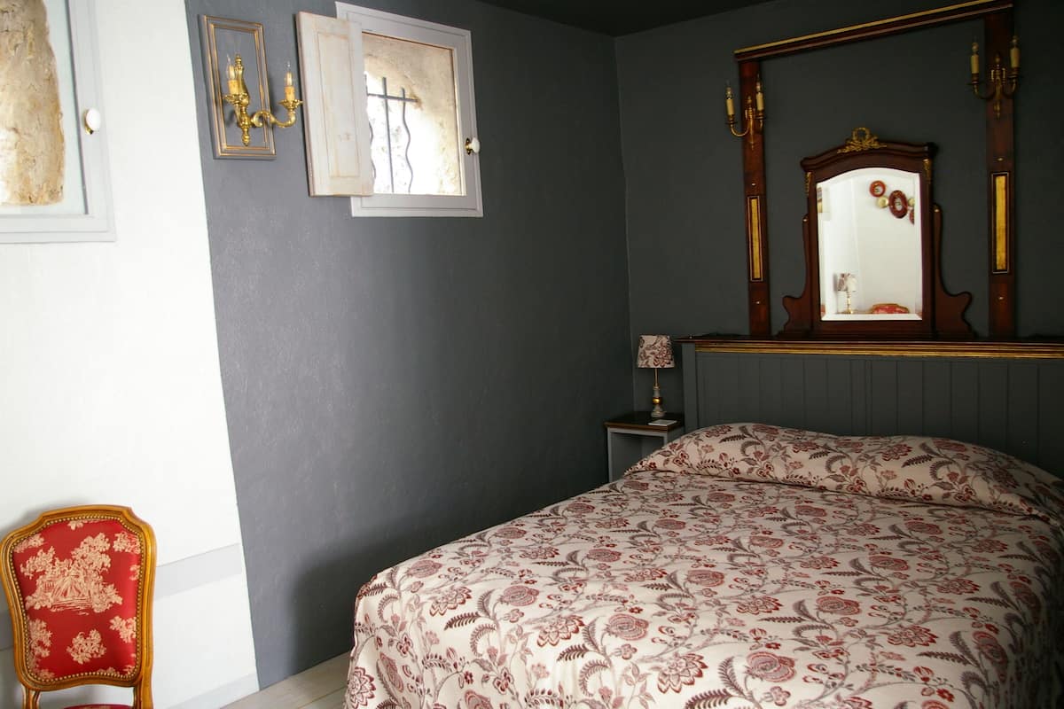 A bedroom with a bed, a red chair, a nightstand with a lamp, a mirror, and framed pictures on the wall.