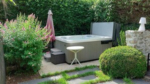 A hot tub in a garden with a table and chairs, surrounded by greenery and a stone wall. - La Source de Bury (Valencisse)