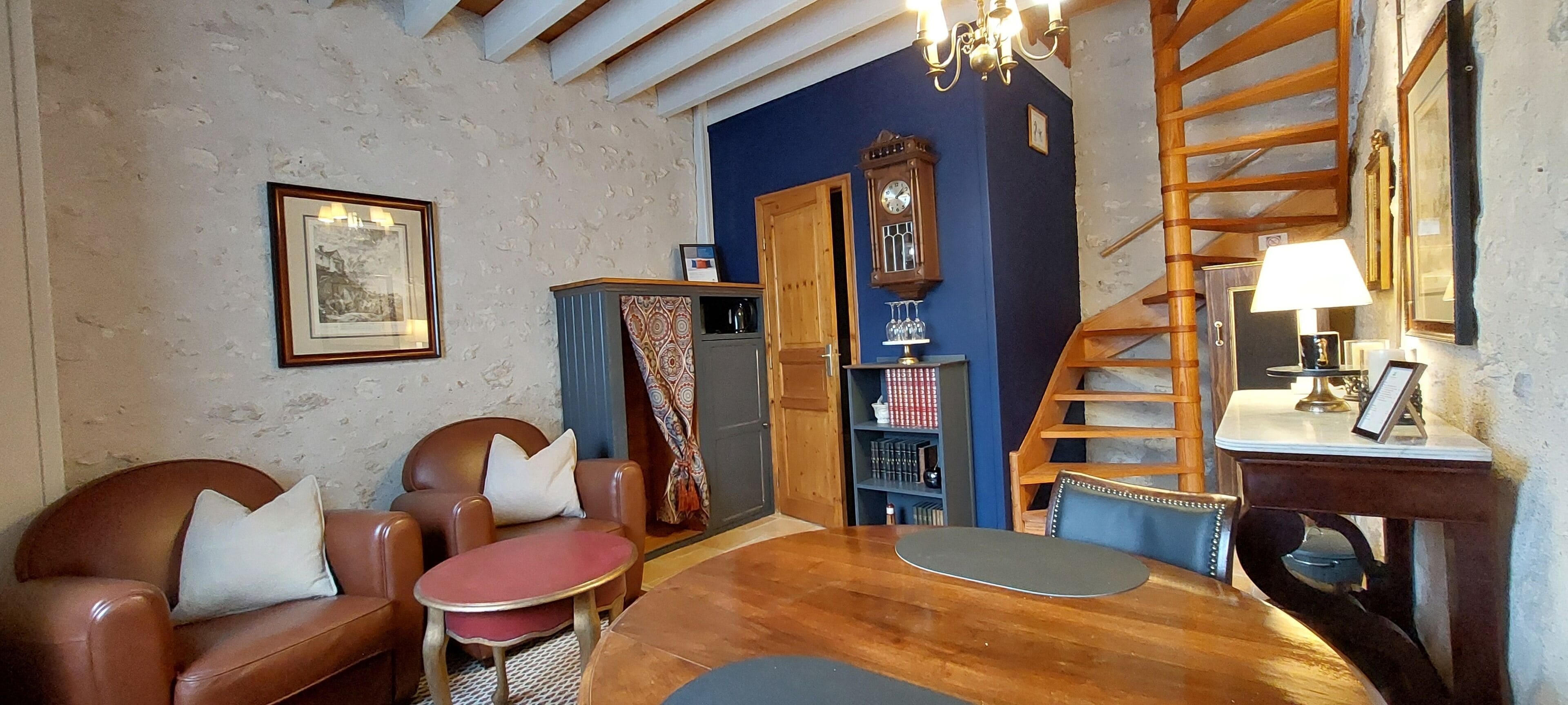 A cozy living room with a wooden table, leather armchairs, a red round table, a blue wall, a wooden staircase, a clock, and a chandelier.