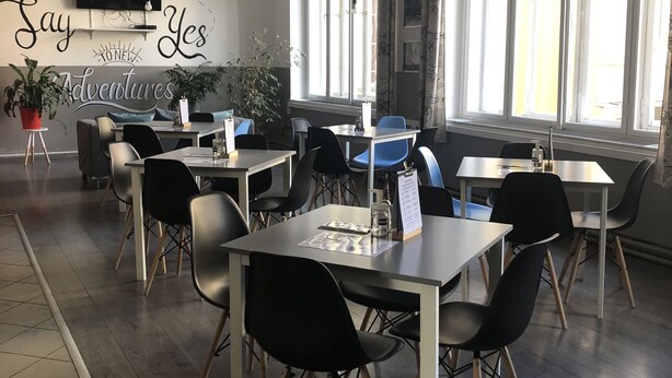 Un café moderno con mesas y sillas, un televisor montado en la pared y un amplio ventanal con cortinas.