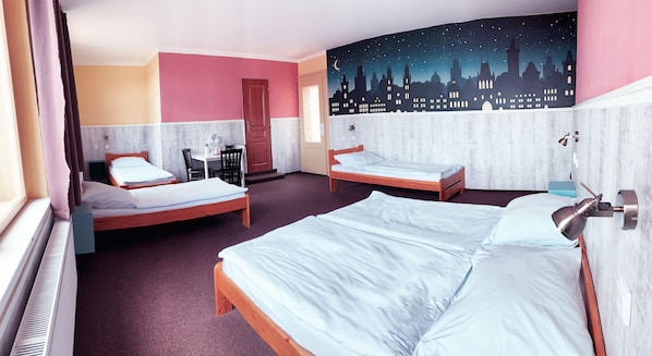 A hotel room with two single beds, a desk with a chair, a small table, a fan, and a night sky mural on the wall.