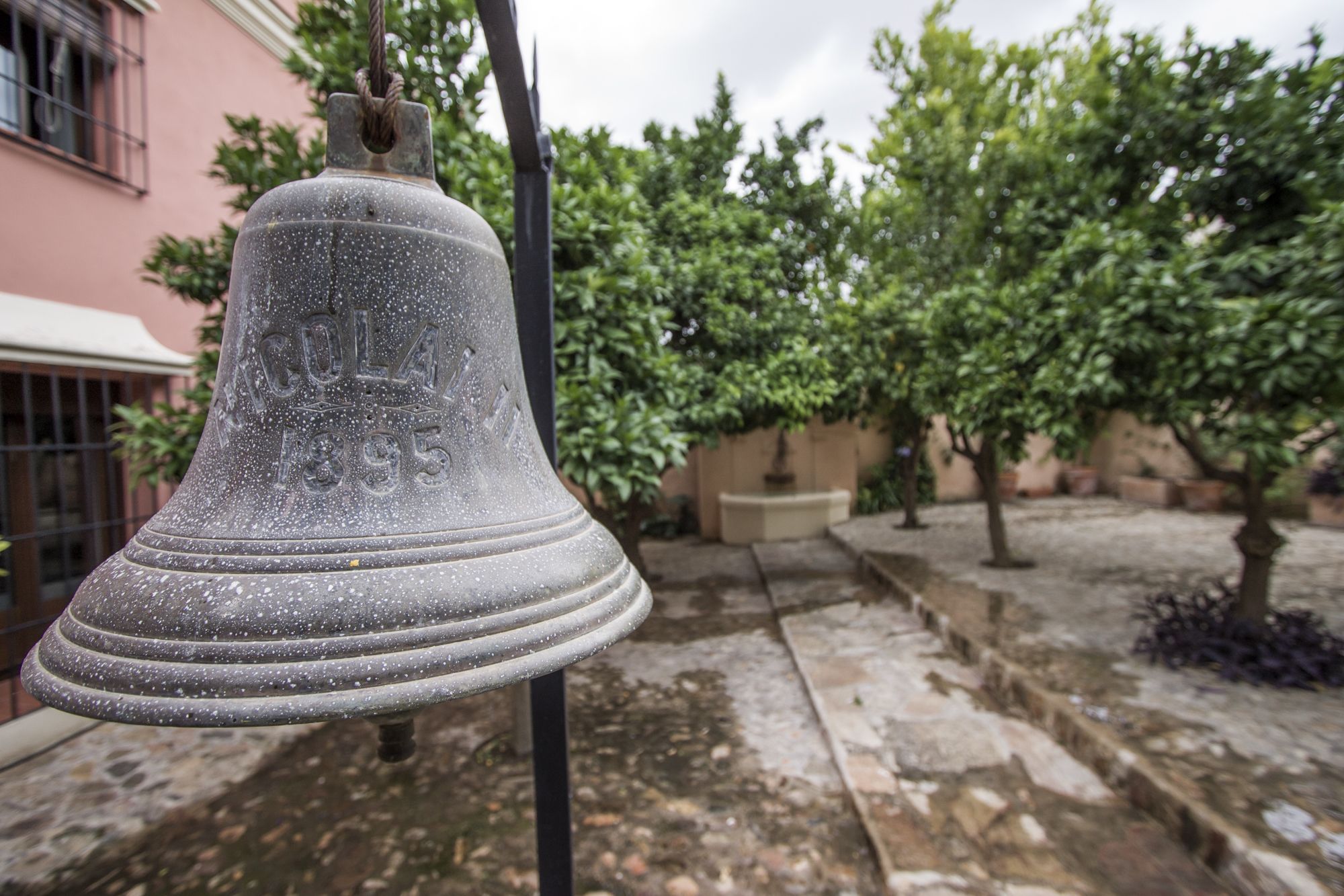 A bell hanging outdoors, likely in a courtyard with a building in the background.