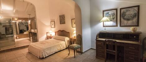 A bedroom with a bed, a desk, and a chair. There are framed pictures on the wall and a lamp on the desk.