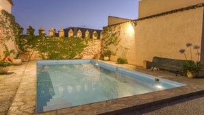 A rectangular swimming pool with a stone border, surrounded by a stone patio. There are potted plants and a bench along the poolside. - Finca Villa Juan (Ribera del Fresno)
